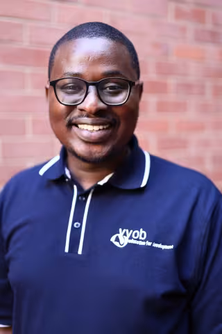 Cristopher smiling in a navy polo shirt stands against a brick wall. His expression is warm and friendly.