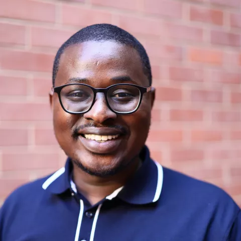 Cristopher smiling in a navy polo shirt stands against a brick wall. His expression is warm and friendly.