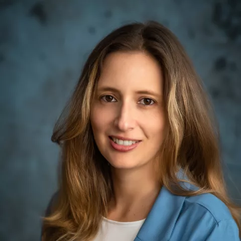 Claudia smiles softly looking into the camera. She wears a light blue blazer over a white shirt. The background is a mottled blue.