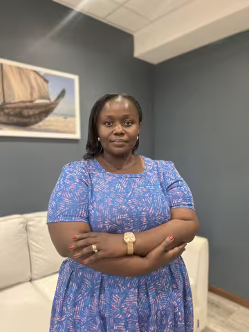 Diana is wearing a blue patterned dress. She stands confidently with arms crossed in a room with gray walls. A boat painting hangs in the background.