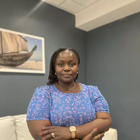 Diana is wearing a blue patterned dress. She stands confidently with arms crossed in a room with gray walls. A boat painting hangs in the background.