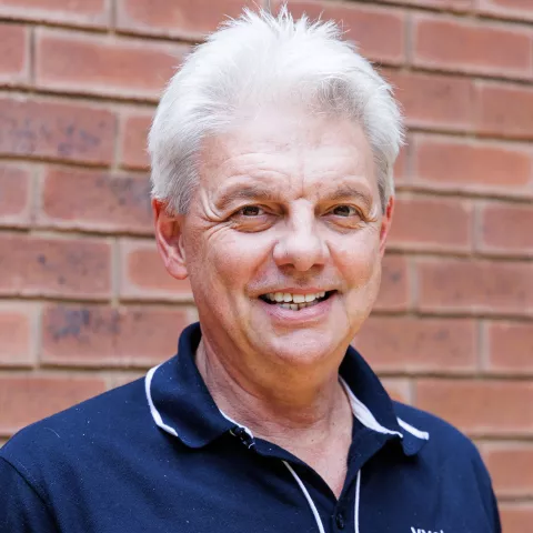 Wilfried smiling in a navy polo shirt stands against a brick wall. His expression is warm and friendly.