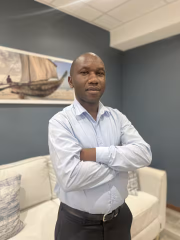 Felix is wearing a light blue shirt and stands confidently with arms crossed in a living room. Behind him is a painting of a boat.