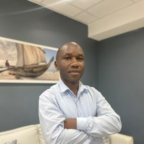 Felix is wearing a light blue shirt and stands confidently with arms crossed in a living room. Behind him is a painting of a boat.