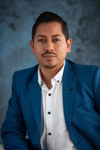 Fernando in a blue suit and white shirt poses confidently against a textured blue-gray backdrop. 