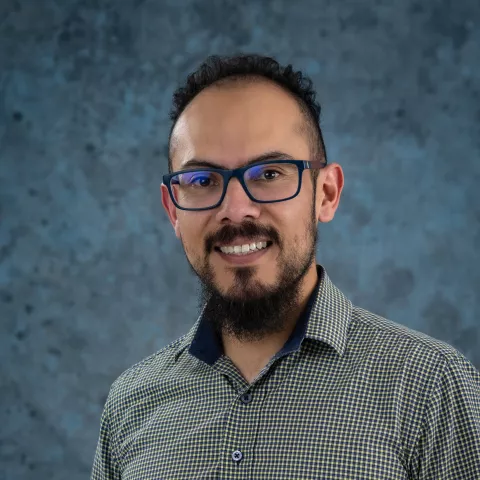 Francisco smiles at the camera. He wears glasses and a green and blue squared shirt. The background is a malted blue backdrop.