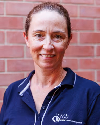 Hanne smiling in a navy polo shirt stands against a brick wall. Her expression is warm and friendly.