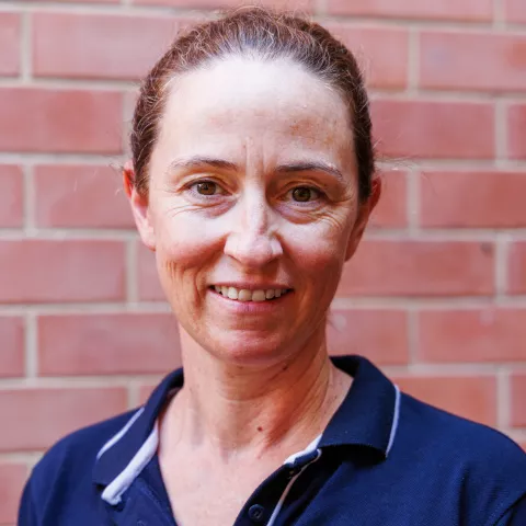 Hanne smiling in a navy polo shirt stands against a brick wall. Her expression is warm and friendly.