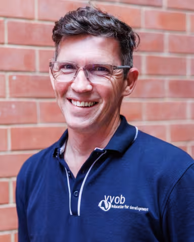 Henre smiling in a navy polo shirt stands against a brick wall. His expression is warm and friendly.
