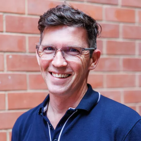 Henre smiling in a navy polo shirt stands against a brick wall. His expression is warm and friendly.