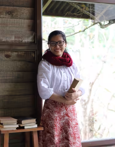 MaiAnh standing indoors by a window, holding a book. She wears glasses, a red scarf, a white blouse, and a patterned skirt. 