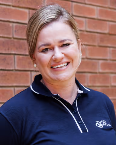 Ise-Lu smiling in a navy polo shirt stands against a brick wall. Her expression is warm and friendly.