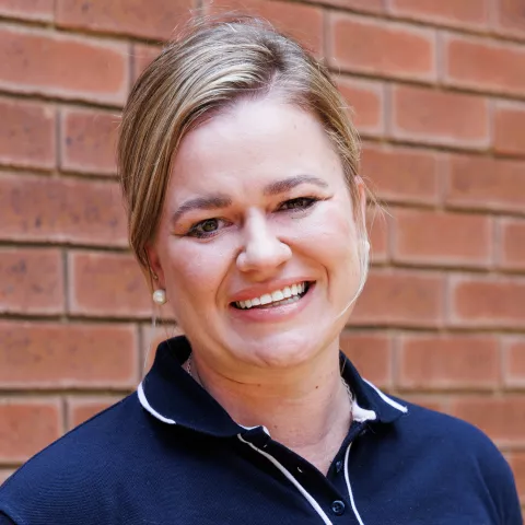 Ise-Lu smiling in a navy polo shirt stands against a brick wall. Her expression is warm and friendly.
