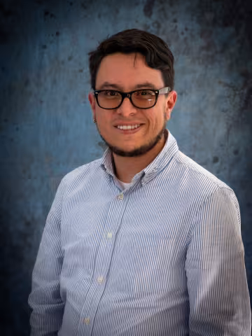 Jose smiles at the camera. He wears glasses and a blue shirt. The background is a malted blue backdrop.