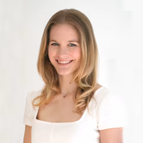 Kelsey smiles warmly at the camera. She is wearing a white shirt and standing in front of a white wall. 