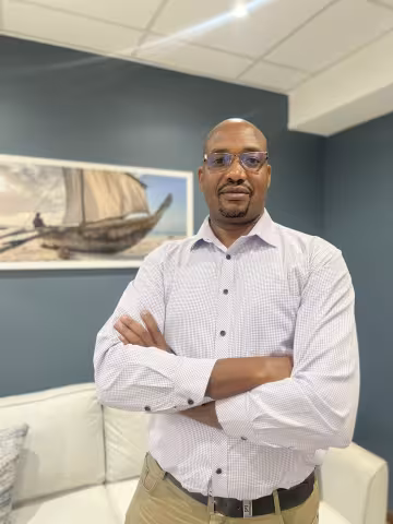  Kube is wearing a light blue shirt and glasses. He stands confidently with arms crossed in a living room. Behind him is a painting of a boat 