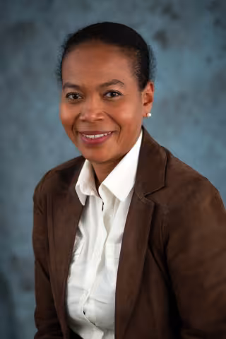 Lepida sits smiling at the camera. She wears a brown blazer over a white shirt.