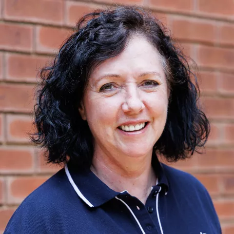 Laetitia smiling in a navy polo shirt stands against a brick wall. Her expression is warm and friendly.