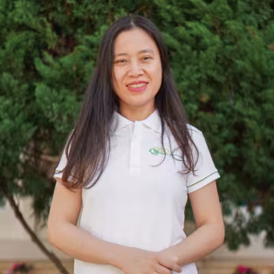 Lieu smiles warmly, wearing a white polo shirt. She stands against a backdrop of lush green foliage.