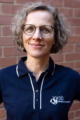 Lieve smiling in a navy polo shirt stands against a brick wall. Her expression is warm and friendly.