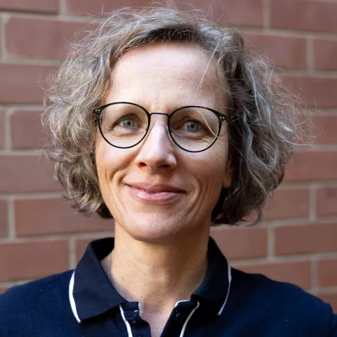 Lieve smiling in a navy polo shirt stands against a brick wall. Her expression is warm and friendly.