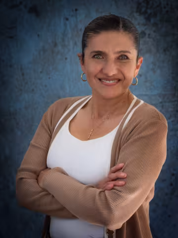 Monica smiles softly looking into the camera. She wears a light brown cardigan over a white shirt. The background is a mottled blue wall.