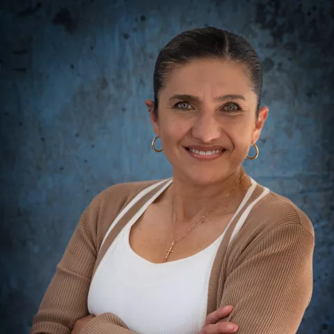 Monica smiles softly looking into the camera. She wears a light brown cardigan over a white shirt. The background is a mottled blue wall.