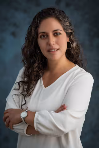Soledad smiles softly looking into the camera. She wears a white shirt. The background is a mottled blue.