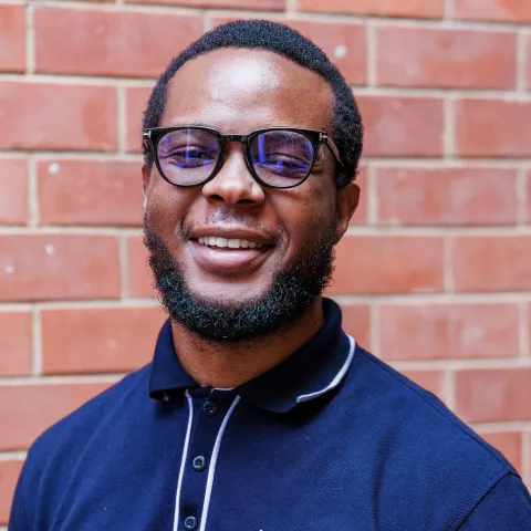 Mawande smiling in a navy polo shirt stands against a brick wall. His expression is warm and friendly.