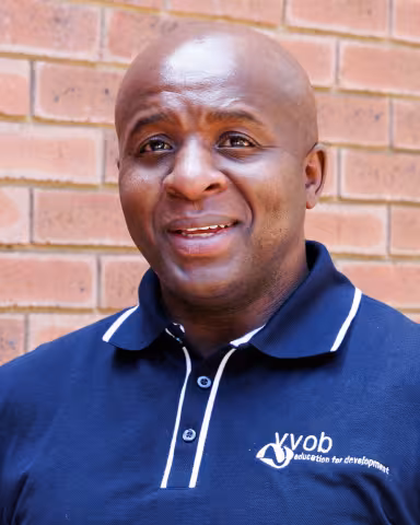 Nkululeko smiling in a navy polo shirt stands against a brick wall. His expression is warm and friendly.