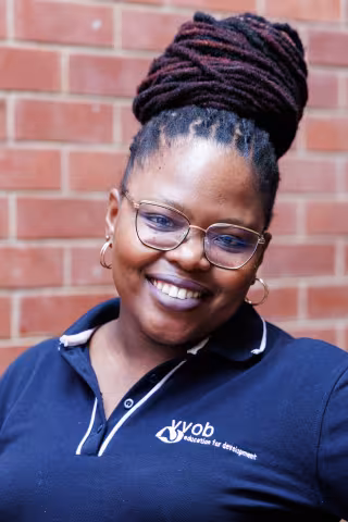 Nondyebo smiling in a navy polo shirt stands against a brick wall. Her expression is warm and friendly.