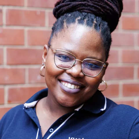 Nondyebo smiling in a navy polo shirt stands against a brick wall. Her expression is warm and friendly.