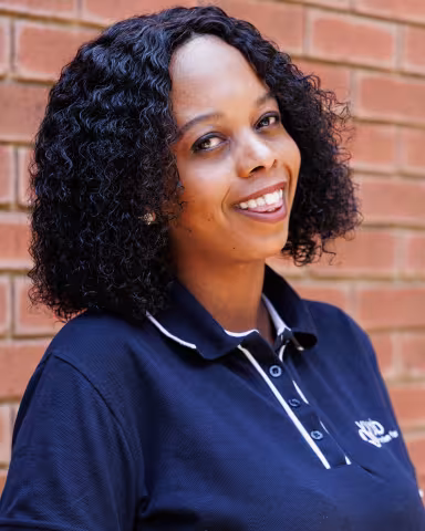 Nosizwe smiling in a navy polo shirt stands against a brick wall. Her expression is warm and friendly.