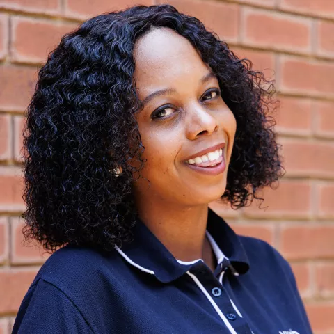 Nosizwe smiling in a navy polo shirt stands against a brick wall. Her expression is warm and friendly.