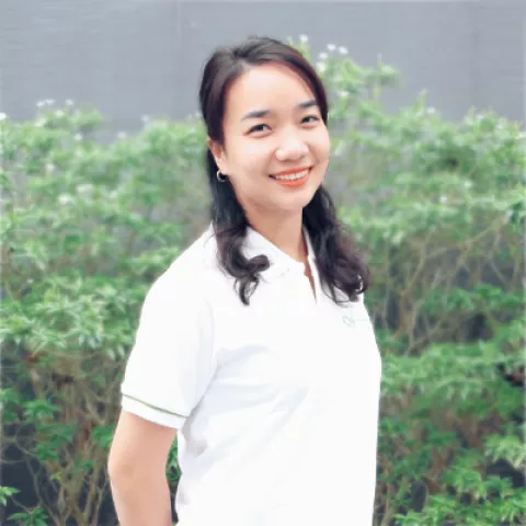 Chau smiles warmly, wearing a white polo shirt. She stands against a backdrop of lush green foliage.