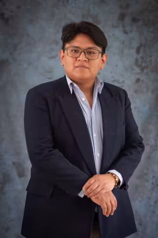Roberto in a navy blazer and light blue shirt stands confidently against a textured gray background. He wears glasses and have short dark hair.