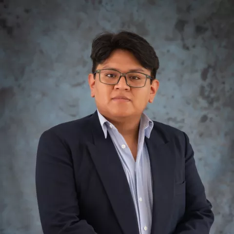 Roberto in a navy blazer and light blue shirt stands confidently against a textured gray background. He wears glasses and have short dark hair.