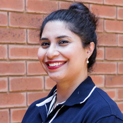 Sahar smiling in a navy polo shirt stands against a brick wall. Her expression is warm and friendly.