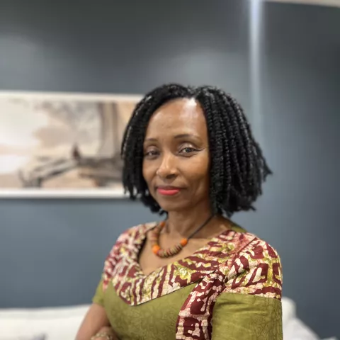 Sarina is wearing a colorful patterned dress and orange necklace, stands confidently with folded arms. The background is a room with blue walls.
