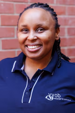 Sisonke smiling in a navy polo shirt stands against a brick wall. Her expression is warm and friendly.