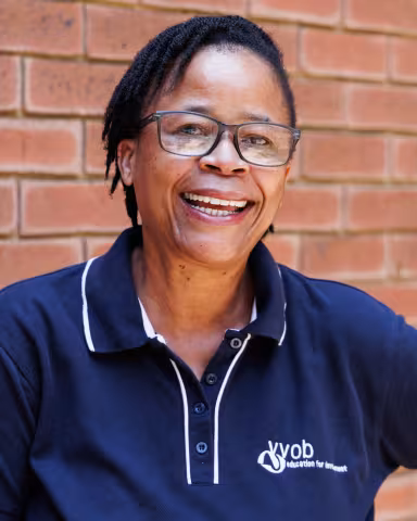 Sylvia smiling in a navy polo shirt stands against a brick wall. Her expression is warm and friendly.