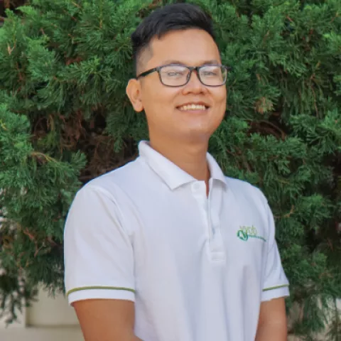 Thanh smiles warmly, wearing a white VVOB polo shirt. He stands against a backdrop of lush green foliage.