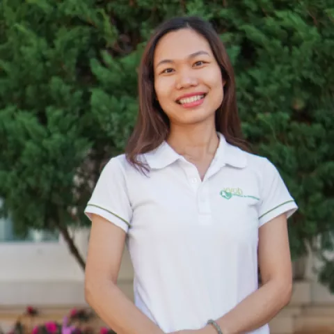 Thanh smiles warmly, wearing a white polo shirt. She stands against a backdrop of lush green foliage.