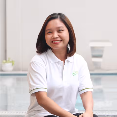 Thinh smiles warmly, wearing a white polo shirt. She sits against a white background with a pot of plant on the side. 