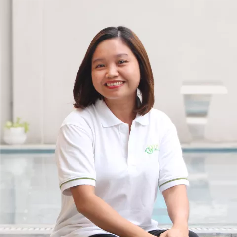 Thinh smiles warmly, wearing a white polo shirt. She sits against a white background with a pot of plant on the side. 