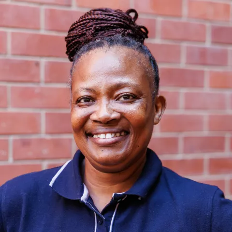Thobile smiling in a navy polo shirt stands against a brick wall. Her expression is warm and friendly.