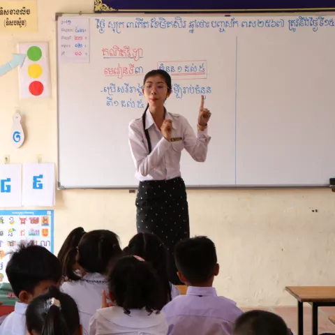 Teacher in a classroom writes on a whiteboard while students sit attentively.