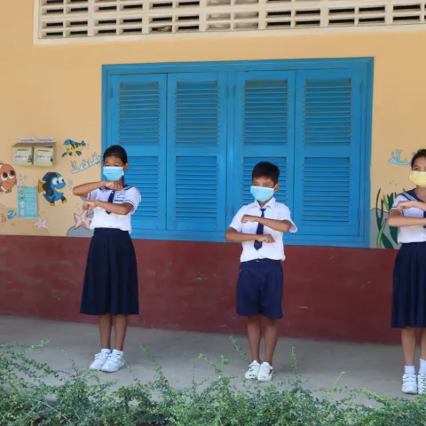 Four students in uniforms and masks standing outside and creating an equal sign with their arms. 