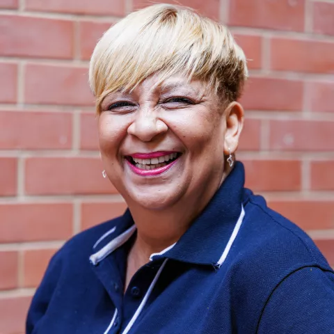 Vanessa smiling in a navy polo shirt stands against a brick wall. Her expression is warm and friendly.
