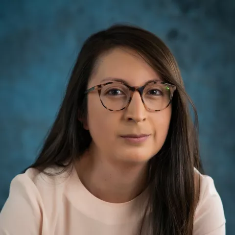 Xiomari smiles softly at the camera. She wears glasses and a beige shirt. The background is a malted blue backdrop.